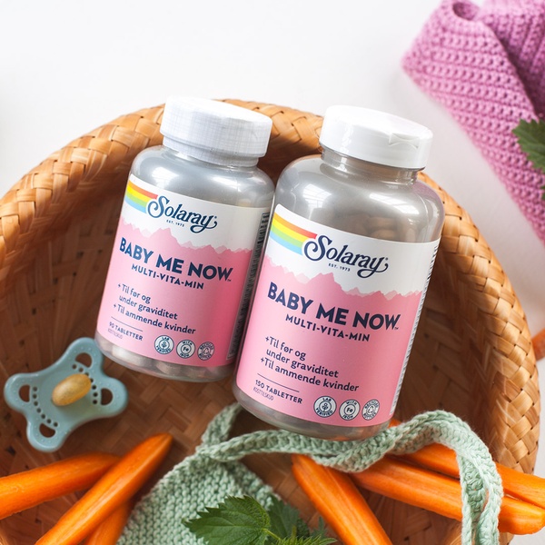 This ad creative displays two bottles of Baby Me Now multi-vitamin supplements placed inside a woven basket with natural elements like carrots and crochet accessories.